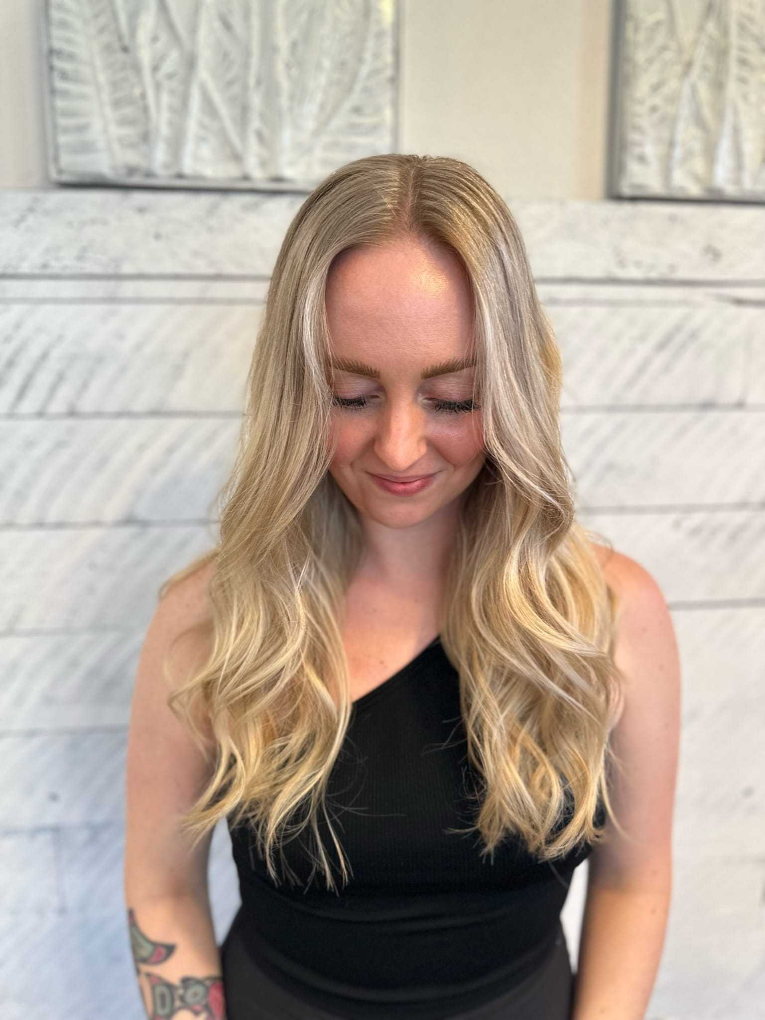 Woman with long, wavy blonde hair smiling and wearing a sleeveless black top, in front of a light background.